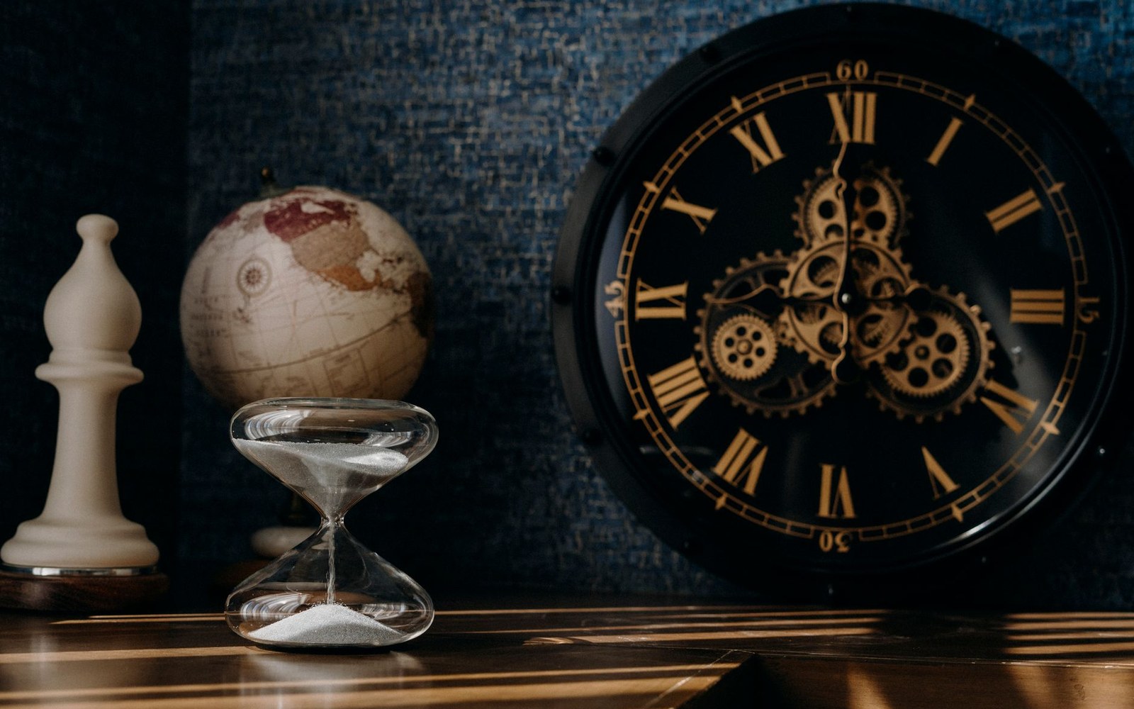 A gear-faced wall clock, hourglass, chess bishop and globe arranged on a wooden desk